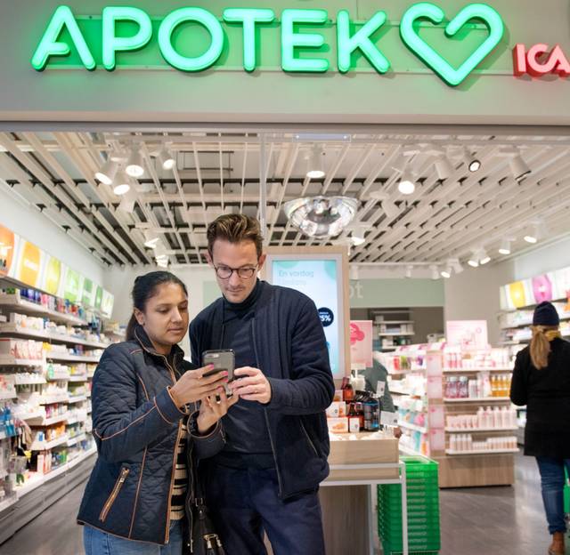 A man and a woman standing inside a brightly lit pharmacy, looking at a smartphone together. In the