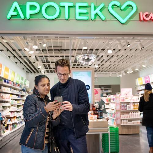 A man and a woman standing inside a brightly lit pharmacy, looking at a smartphone together. In the